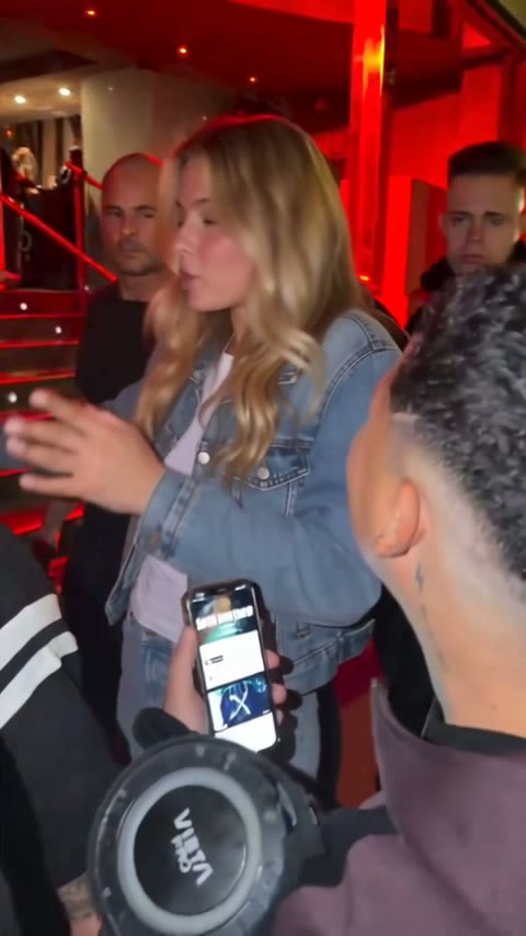 A young woman with long wavy blonde hair in a denim jacket rapping at the red-lit entrance